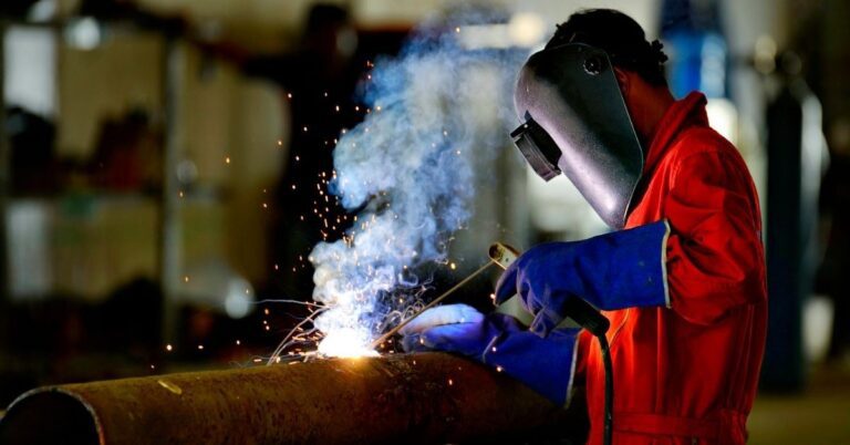 A person wearing a red protective suit, blue gloves, and a welding helmet is welding a large rusty metal pipe, with bright sparks and smoke visible at the welding point. The background is blurred, focusing attention on the welding activity.