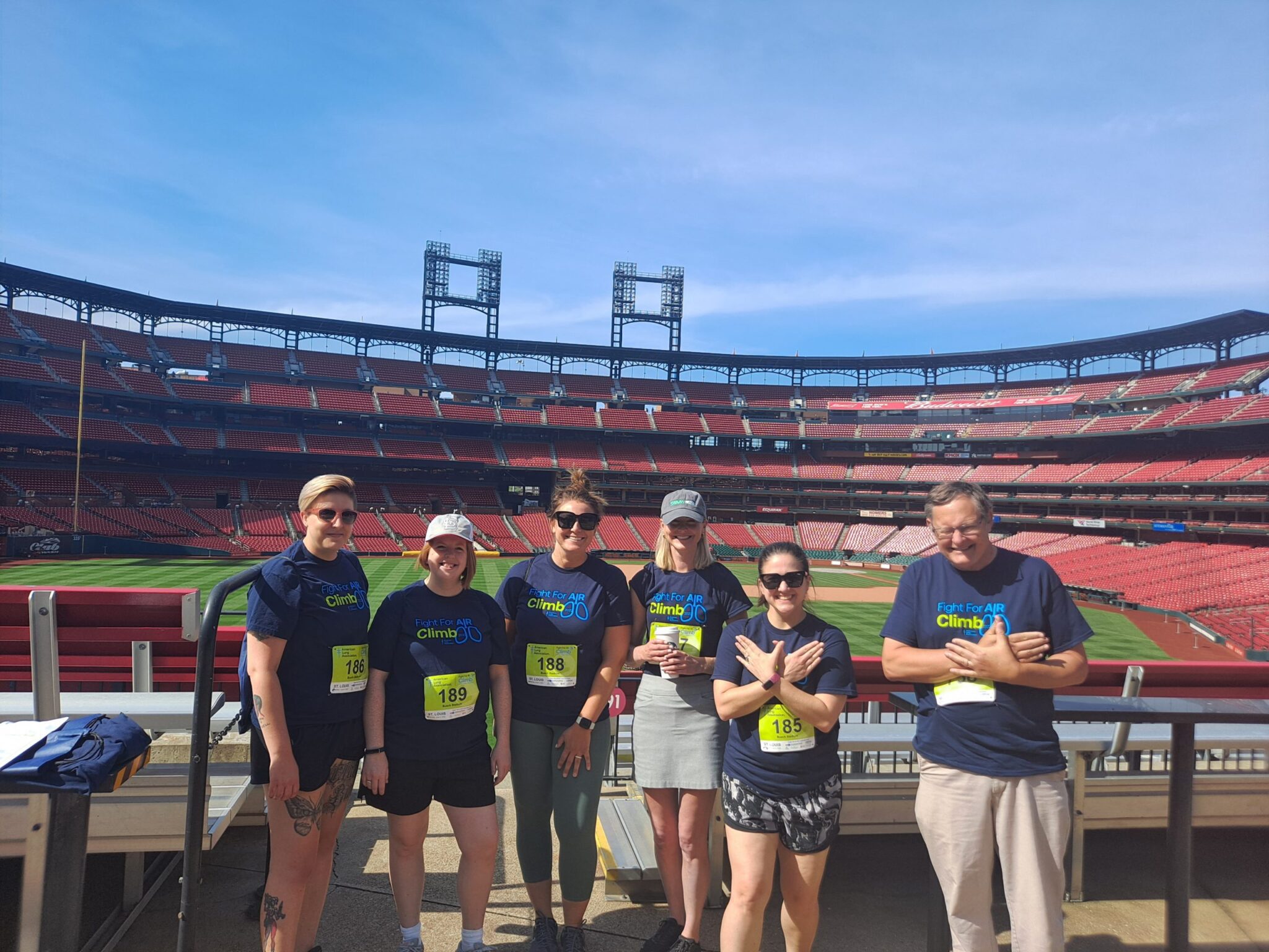 Six people wearing matching navy blue "Fight For Air Climb 30" t-shirts and yellow race bibs stand in front of a baseball field inside a large stadium with red seats. The group is posing with some crossing their arms in front of their chests. The sky is clear and blue.