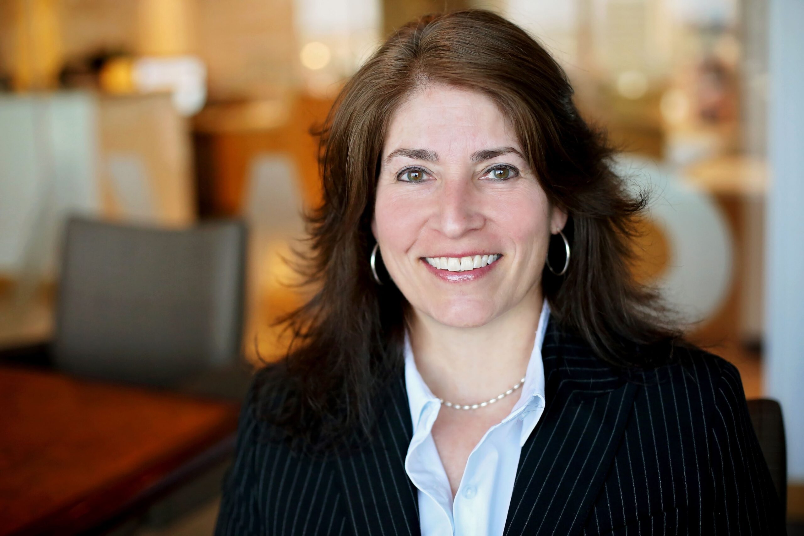 Woman with shoulder-length brown hair wearing a black pinstripe blazer, light blue shirt, pearl necklace, and hoop earrings, smiling in an office setting with blurred background.