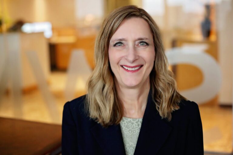 Woman with shoulder-length blonde hair, wearing a dark blazer over a light lace top, smiling in a softly lit indoor setting with blurred background elements.