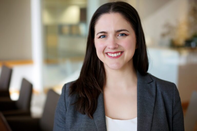 A smiling woman with long dark hair wearing a gray blazer over a white top, standing in a softly blurred indoor setting with chairs and light-colored walls in the background.