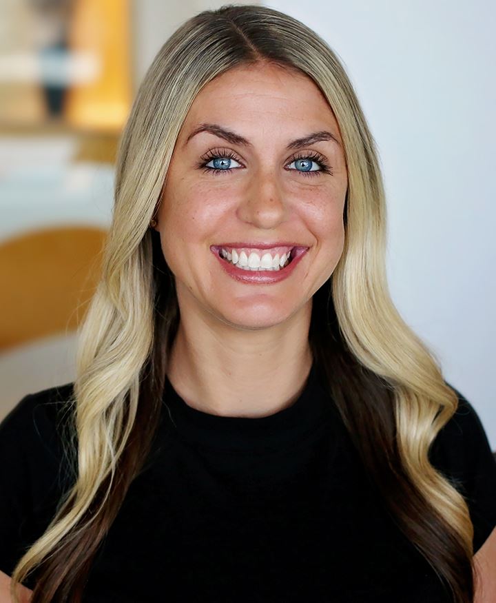 Smiling woman with long, wavy blonde hair featuring darker roots, bright blue eyes, and wearing a black top, posed against a blurred indoor background.