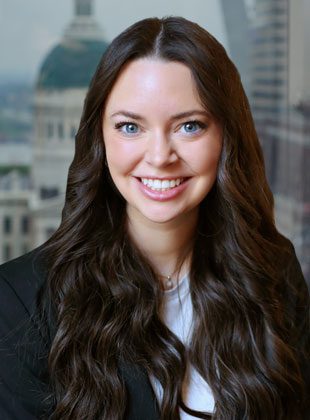 Young woman with long, wavy dark brown hair and blue eyes, smiling. She is wearing a black blazer over a white top, with a blurred cityscape background featuring a domed building.