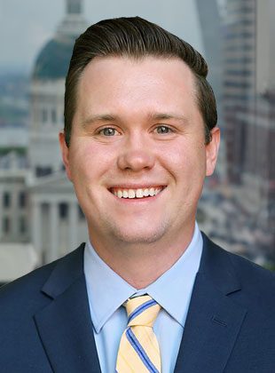 A smiling man with neatly combed dark hair wearing a navy blue suit jacket, light blue dress shirt, and a yellow tie with blue diagonal stripes. The background shows a blurred cityscape with a domed building.
