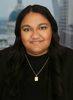 A woman with long, dark hair wearing a black top and black blazer. She has a small nose stud and is wearing a gold necklace with a rectangular pendant. The background shows a blurred cityscape with buildings.