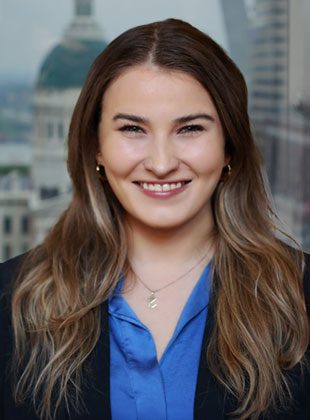 A woman with long, wavy brown hair wearing a blue blouse and a black blazer. She is smiling and wearing small hoop earrings and a delicate necklace. The background shows a blurred cityscape with a domed building.