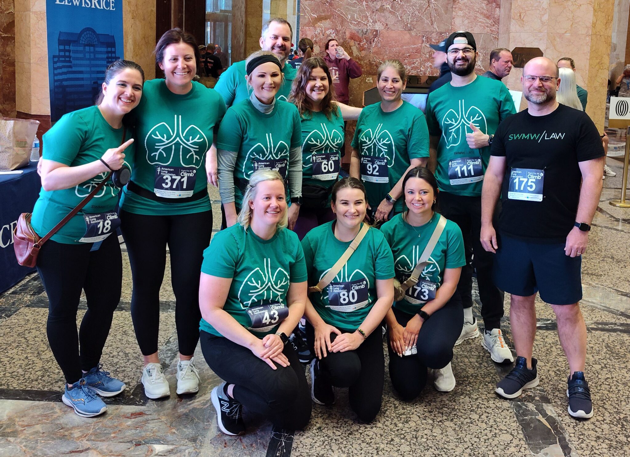 A group of eleven people posing indoors, most wearing matching green t-shirts with a white lung design and race bibs for the American Lung Association Fight for Air Climb event. One person on the right wears a black "SWMW LAW" t-shirt with a race bib. The group is smiling, with some standing and others kneeling on a marble floor. The background shows a marble wall and other people in the distance.
