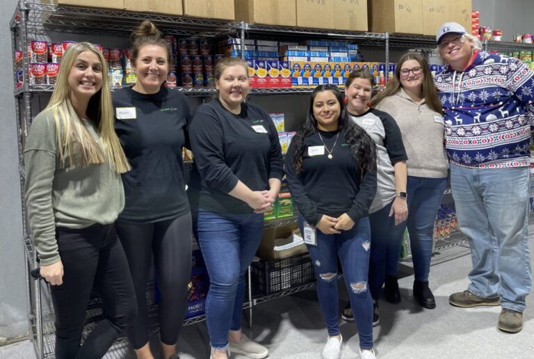 Seven people stand smiling in front of metal shelves stocked with canned goods, boxed pasta, and other food items. They are casually dressed, some wearing name tags, and appear to be in a food pantry or storage area. The group includes six women and one man, who is wearing a festive sweater with reindeer and snowflake patterns and a baseball cap.