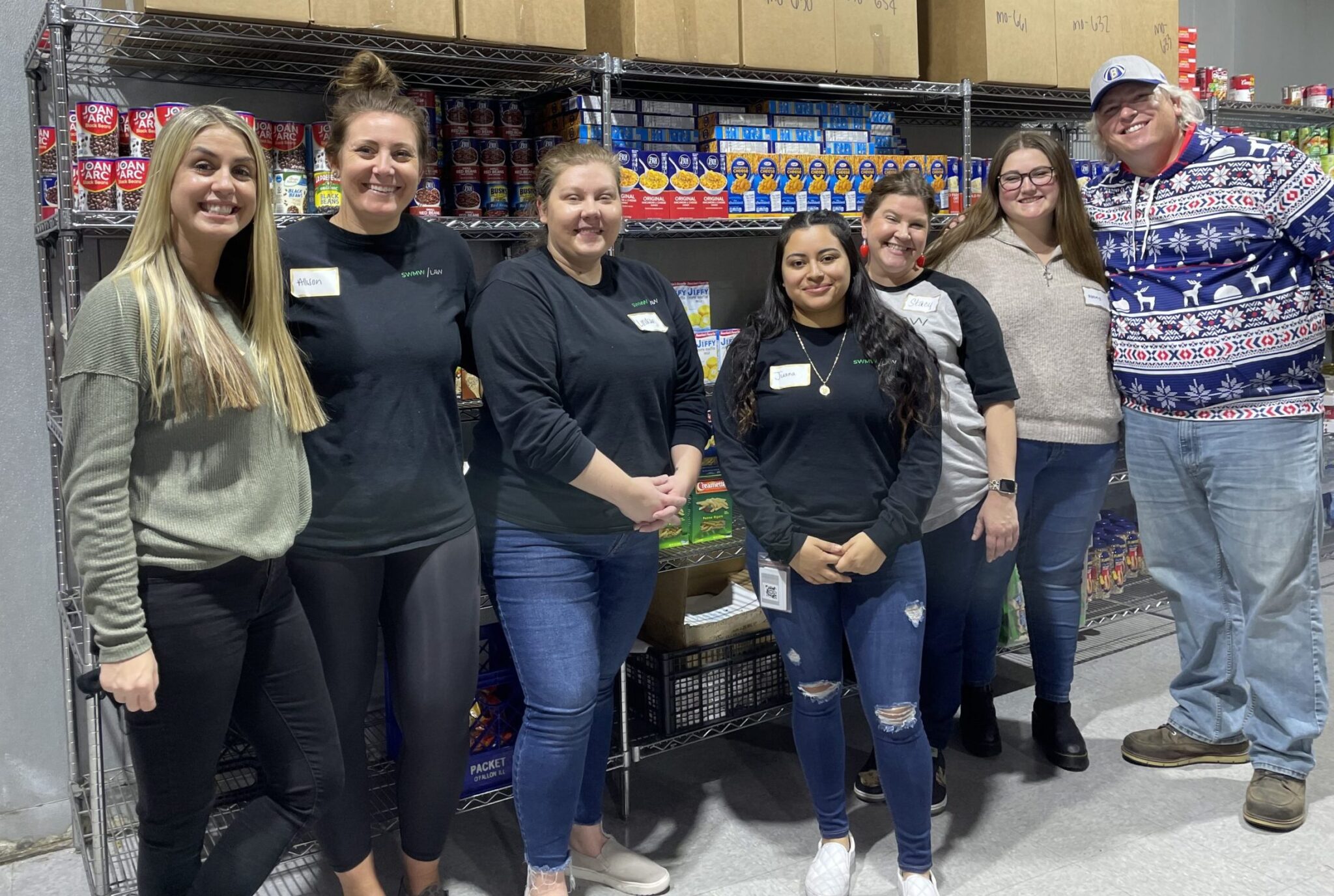Seven people stand smiling in front of metal shelves stocked with canned goods, boxed pasta, and other food items. They are casually dressed, some wearing name tags, and appear to be in a food pantry or storage area. The group includes six women and one man, who is wearing a festive sweater with reindeer and snowflake patterns and a baseball cap.