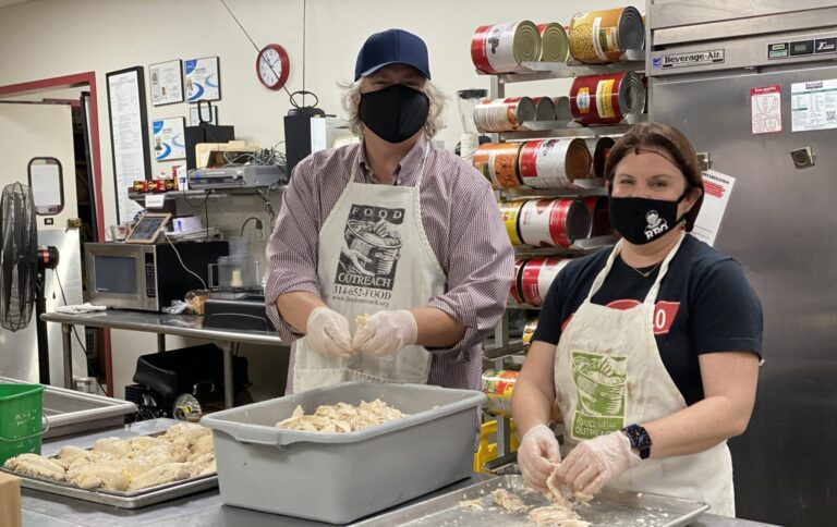 Two people wearing aprons and gloves are preparing shredded chicken in a kitchen. Both are wearing face masks, and behind them are large cans stacked on shelves and a stainless steel refrigerator. The person on the left is wearing a blue cap and a checkered shirt, while the person on the right is wearing a black t-shirt and a smartwatch.