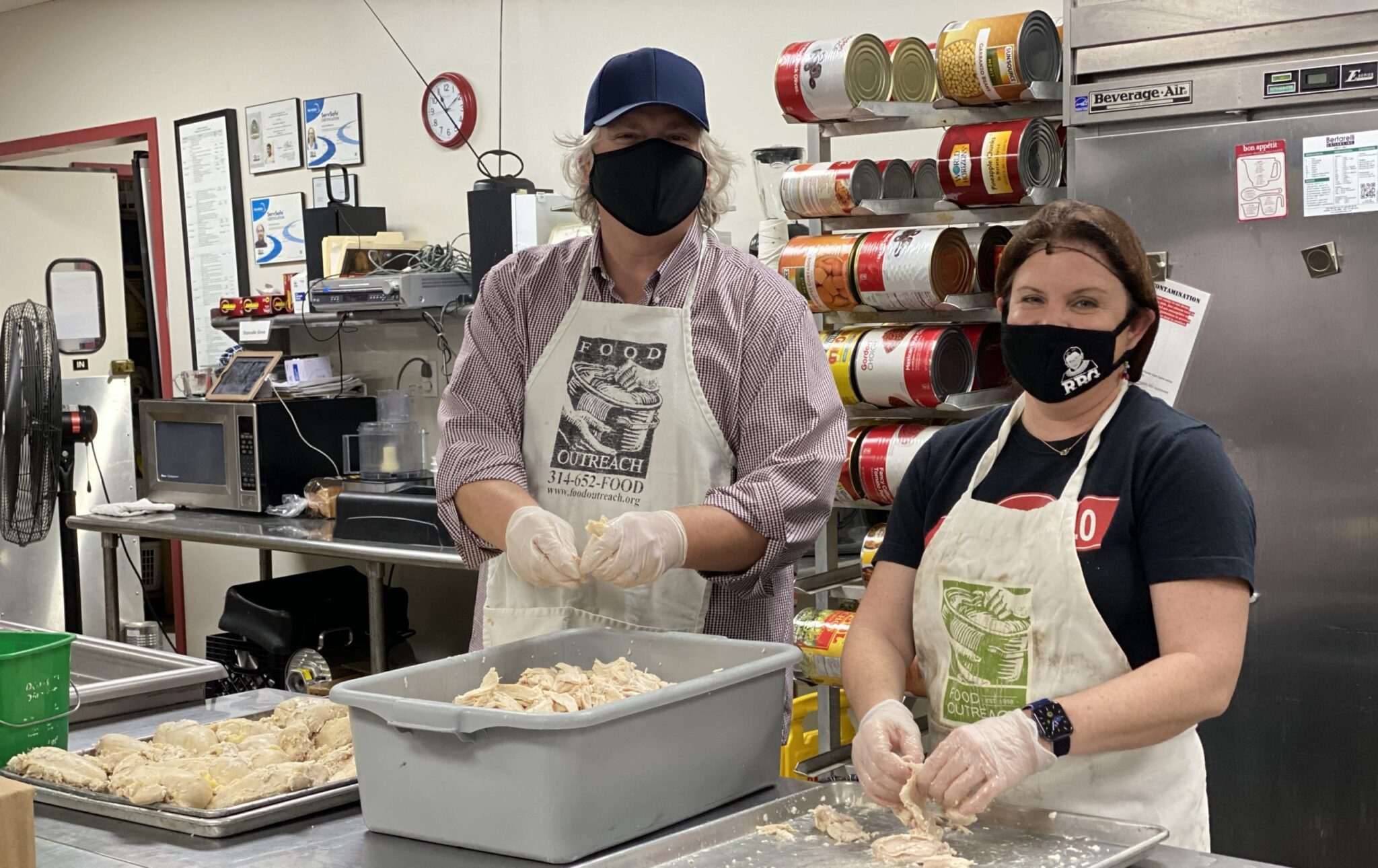 Two people wearing aprons and gloves are preparing shredded chicken in a kitchen. Both are wearing face masks, and behind them are large cans stacked on shelves and a stainless steel refrigerator. The person on the left is wearing a blue cap and a checkered shirt, while the person on the right is wearing a black t-shirt and a smartwatch.