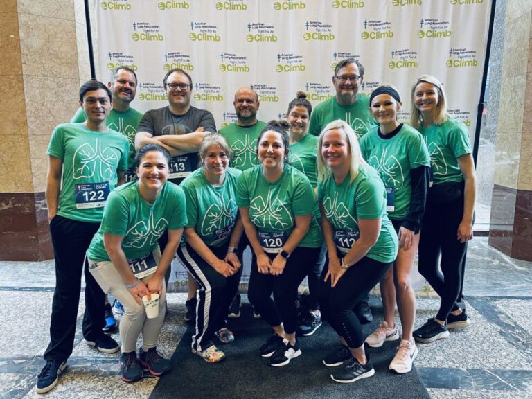 A group of thirteen people wearing matching green t-shirts with a white lung design, posing indoors in front of a backdrop featuring the American Lung Association and Fight For Air Climb logos. Most are wearing black or dark pants and athletic shoes, and several have race bibs pinned to their shirts. The group is smiling and appears ready for a fitness or charity event.