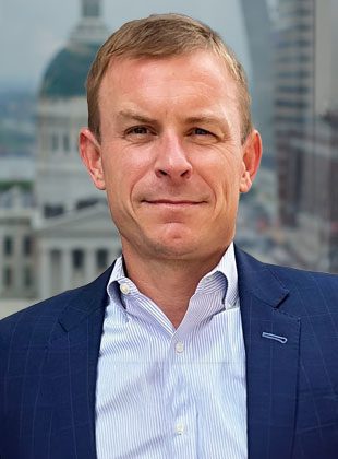 Close-up of a man with short light brown hair wearing a blue suit jacket over a light blue and white striped dress shirt, standing in front of a blurred light-colored building.