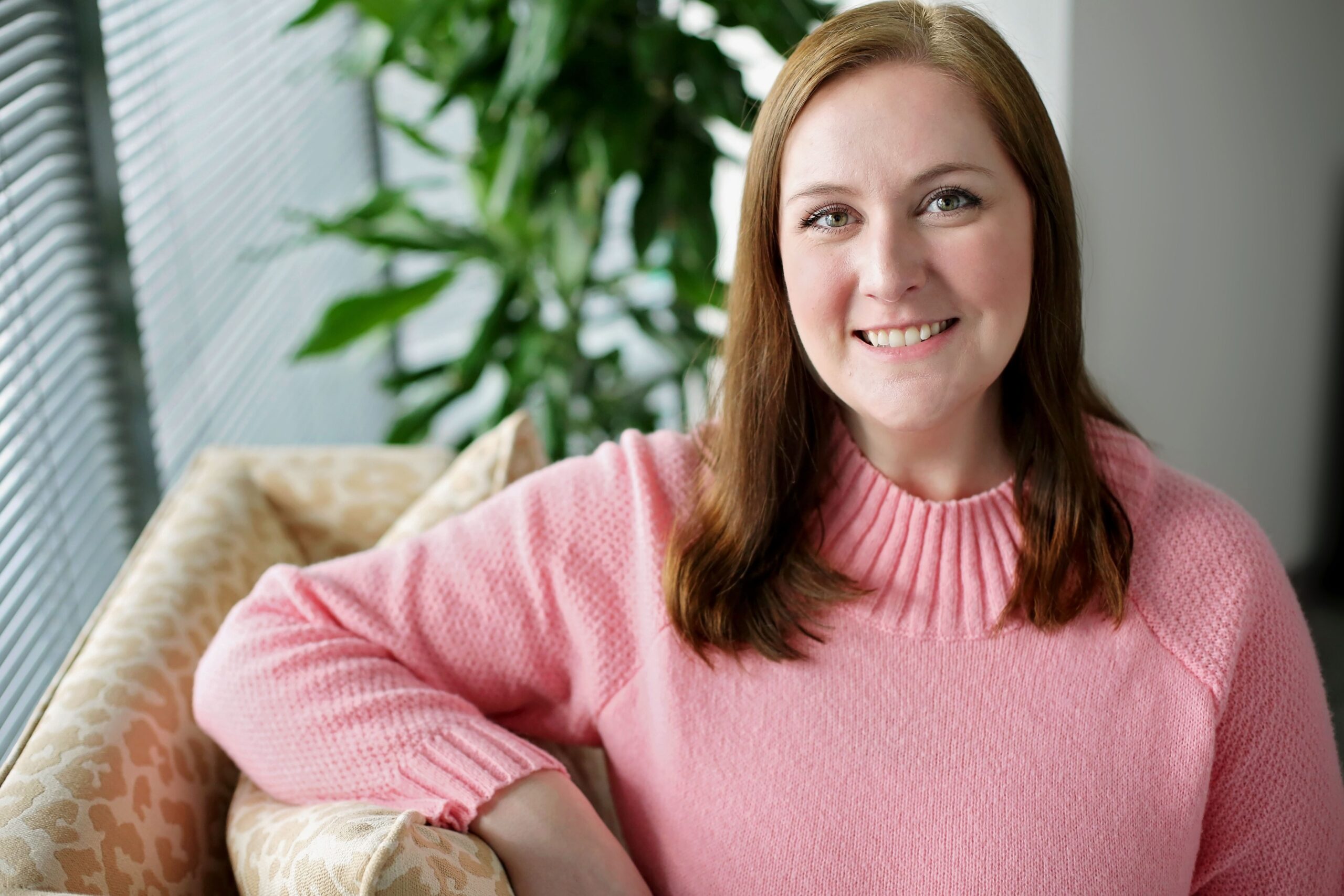 A woman with light skin and straight brown hair is smiling while sitting on a beige patterned couch. She is wearing a pink knit sweater with a ribbed collar and textured sleeves. In the background, there is a green leafy plant and a window with blinds.