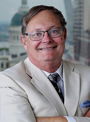 Middle-aged man wearing glasses and a light beige suit with a patterned tie and a blue pocket square, smiling with arms crossed. The background shows an urban cityscape with blurred buildings.
