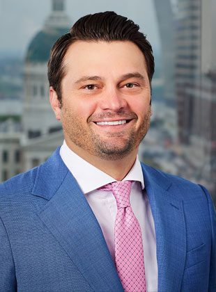 Man with dark hair and light facial stubble wearing a blue suit jacket, white dress shirt, and pink patterned tie, smiling in front of a blurred cityscape background.