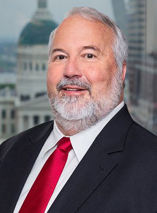 Middle-aged man with gray hair and a beard, wearing a black suit jacket, white dress shirt, and red tie, standing in front of a blurred cityscape background.