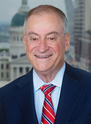 Middle-aged man with short gray hair wearing a dark blue suit, light blue shirt, and red striped tie, smiling in front of a blurred cityscape background featuring a domed building.