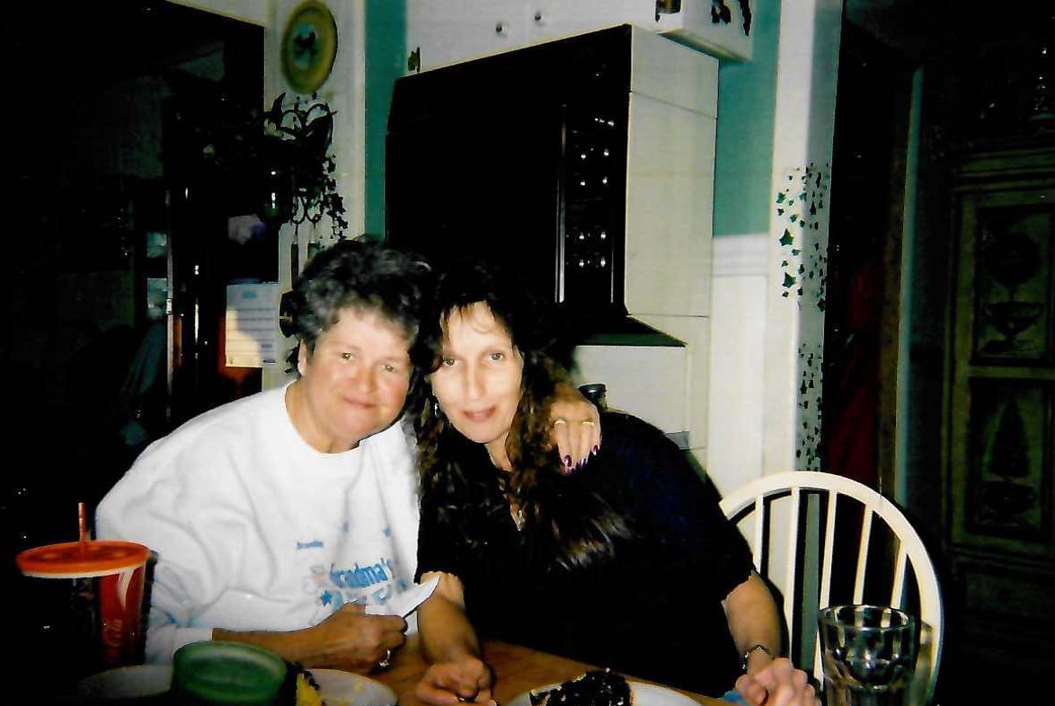 Two women sitting closely together at a table, one wearing a white sweatshirt and the other in a black top. The woman in the white sweatshirt has short curly hair, and the woman in black has long wavy hair. On the table in front of them are plates with food and a red cup with a straw. The background shows a kitchen area with cabinets and a white chair.