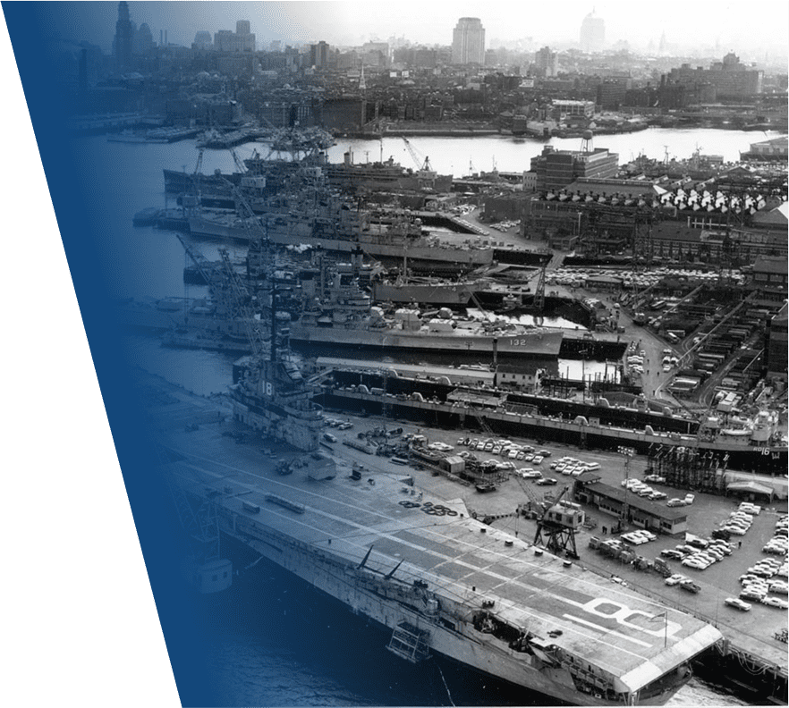 A black and white aerial view of a naval shipyard featuring multiple large warships docked side by side. The foreground shows an aircraft carrier with a visible flight deck marked with the number "8." Behind it, several battleships or cruisers are lined up, with hull numbers "18," "132," and "16" visible. The shipyard is busy with numerous cranes, vehicles, and buildings, and a city skyline is faintly visible in the background through a light haze. The left side of the image has a blue gradient overlay with a green diagonal stripe.