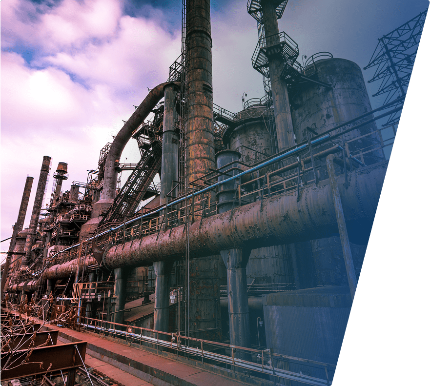 Large industrial factory with tall rusted metal chimneys, pipes, and cylindrical tanks under a partly cloudy sky. The structure appears aged with visible wear and corrosion.