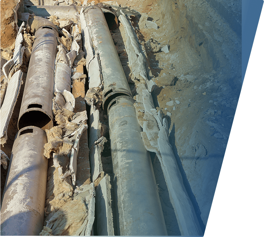 Two large, weathered metal pipes are laid parallel in a trench filled with dirt and broken concrete pieces. The pipes are surrounded by torn insulation material, and the trench walls show exposed soil and rocks. The pipes appear old and possibly part of an underground utility or infrastructure system.