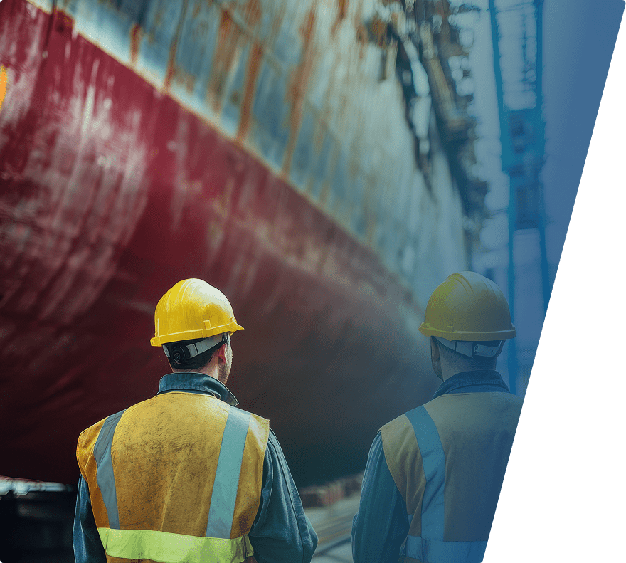 Two construction workers wearing yellow hard hats and reflective vests are standing with their backs to the camera, looking up at the large, rusted hull of a ship in a shipyard. The ship's hull is predominantly red with patches of rust and weathering. The background includes industrial structures and scaffolding.