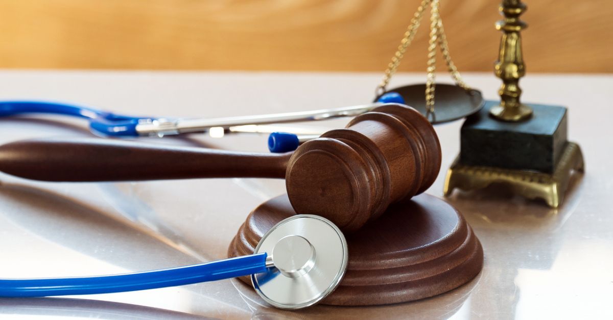 A wooden judge's gavel resting on its sound block, with a blue stethoscope draped over it, and a brass balance scale in the background on a reflective surface.