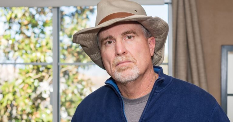 Middle-aged man with light skin and a short gray beard wearing a beige wide-brimmed hat with a brown band, a blue zip-up jacket, and a gray shirt, standing indoors in front of a window with blurred greenery outside.