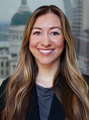 Young woman with long, light brown hair and a nose ring, smiling. She is wearing a dark blazer over a gray top, with a blurred cityscape background.