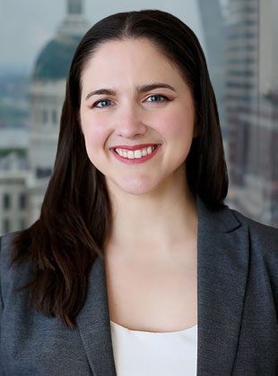 A woman with long dark brown hair, blue eyes, and light skin is smiling. She is wearing a gray blazer over a white top. The background shows a blurred cityscape with buildings.