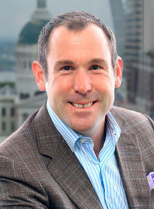 Man with short dark hair wearing a brown plaid blazer over a blue and white striped shirt, smiling, with a purple pocket square, in front of a blurred cityscape background.