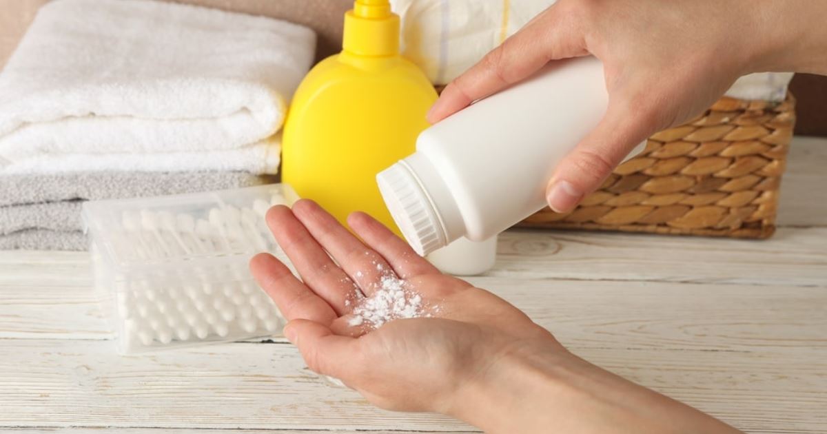 A person is pouring white powder, likely baby powder or talcum powder, from a white bottle onto their open palm. In the background, there are folded towels, a yellow bottle with a pump dispenser, a box of cotton swabs, and a woven basket on a light wooden surface.