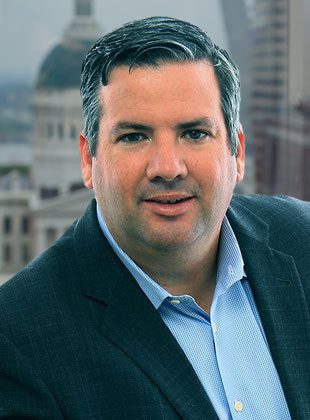 Man with short dark hair wearing a dark blazer over a light blue checkered shirt, posing in front of a blurred cityscape background.