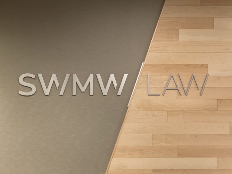 SWMW / LAW sign with metallic letters mounted on a wall that is half covered with a textured gray surface and half with light wood paneling.