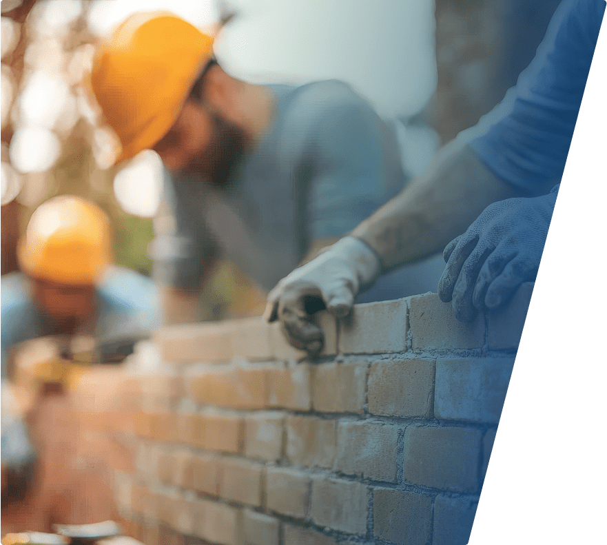 Two construction workers wearing orange helmets and gloves are building a brick wall, focusing on placing and aligning the bricks carefully.
