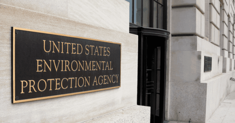 A black and gold plaque mounted on a light stone wall reads "UNITED STATES ENVIRONMENTAL PROTECTION AGENCY." The plaque is near a building entrance with large windows and a revolving door. The building exterior is made of light-colored stone blocks.