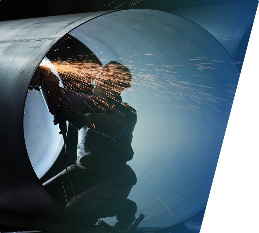 A person wearing protective gear is using a grinder inside a large metal pipe, producing bright sparks. The scene is backlit, creating a silhouette effect against the interior of the pipe.