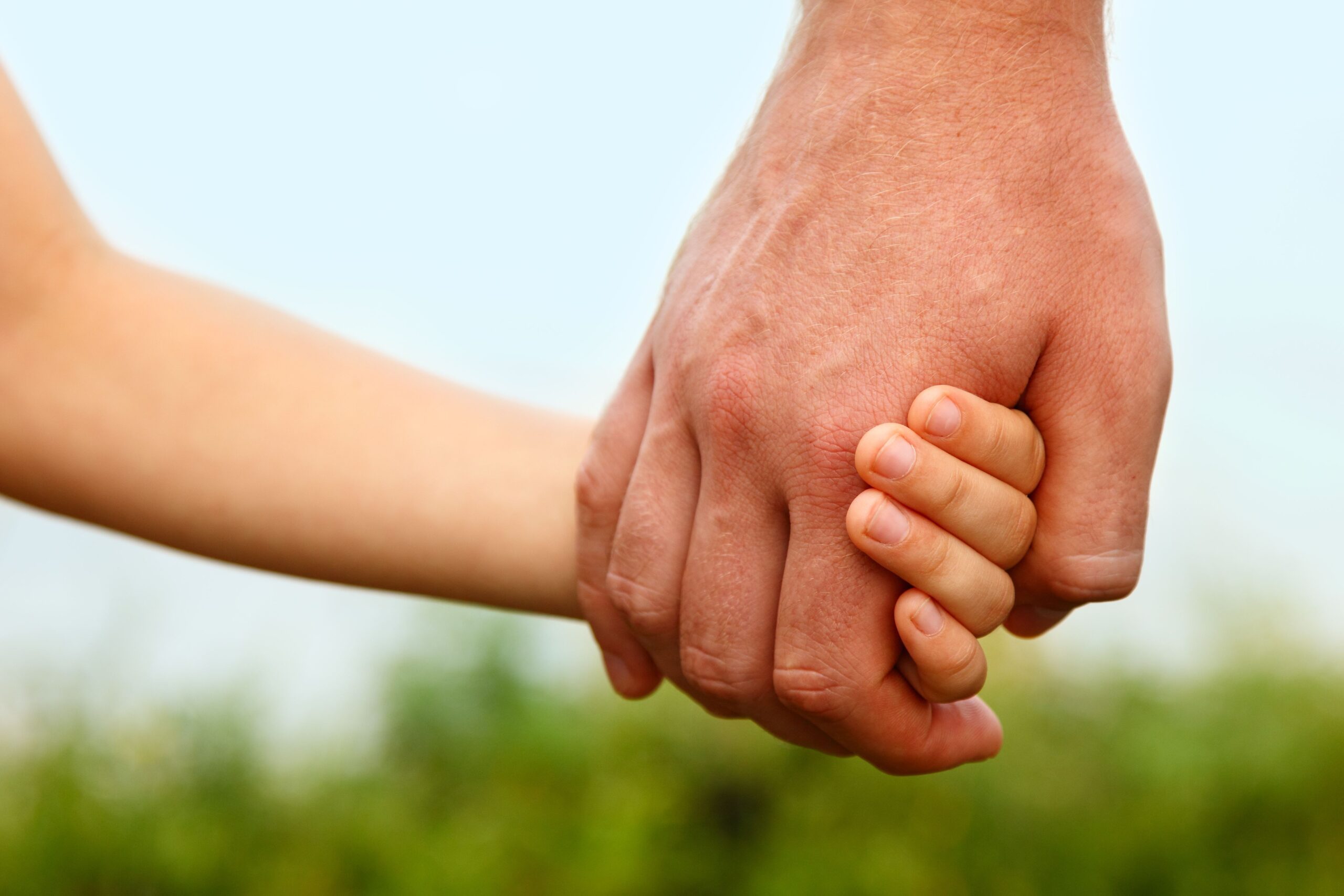 An adult hand holding a child's hand against a blurred outdoor background with greenery and a pale blue sky. The child's fingers are wrapped around the adult's hand.