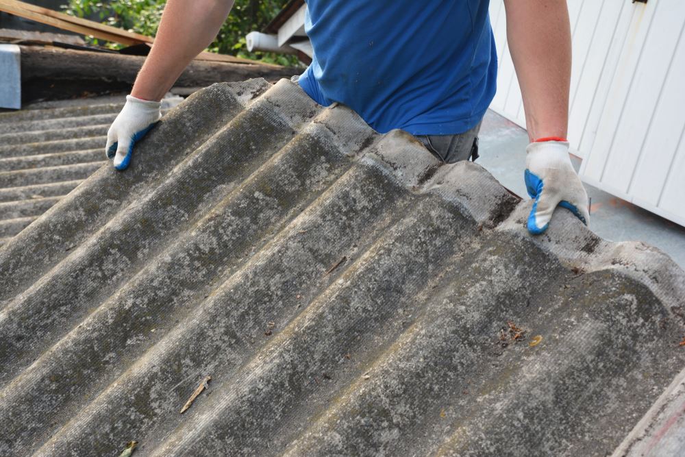 Person wearing a blue shirt and white gloves with blue fingertips holding a large, weathered, corrugated roofing sheet. The sheet appears old and covered with dirt and debris. The background shows part of a roof and a white building wall.
