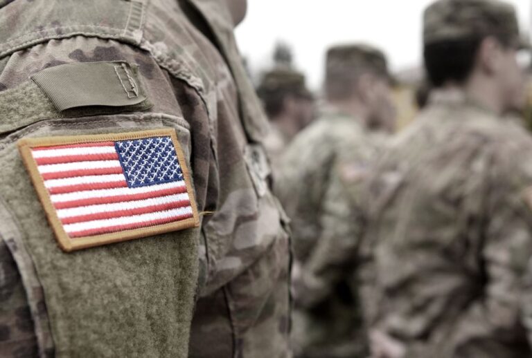 Close-up of a U.S. flag patch on the shoulder of a soldier wearing camouflage uniform, with other soldiers in similar uniforms blurred in the background.