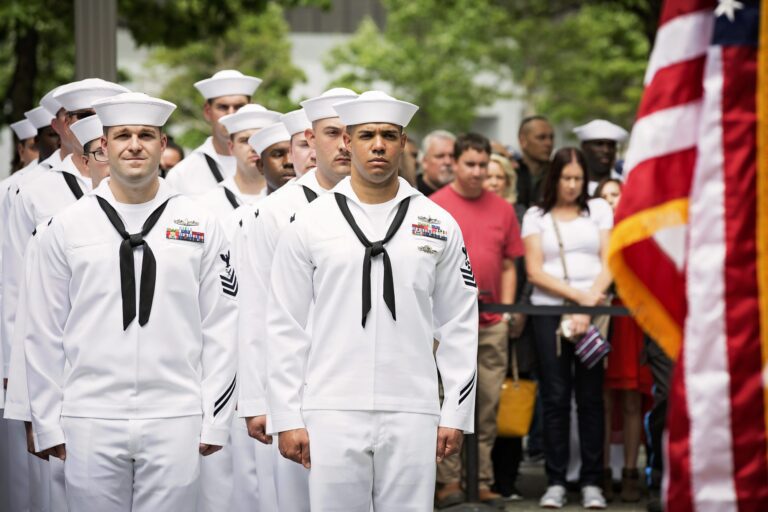 A group of U.S. Navy sailors in white dress uniforms and white sailor hats stand in formation outdoors. They have black neckerchiefs and various service ribbons on their chests. Behind them, a crowd of civilians watches, and an American flag is partially visible on the right side of the image. Trees and a building are in the background.