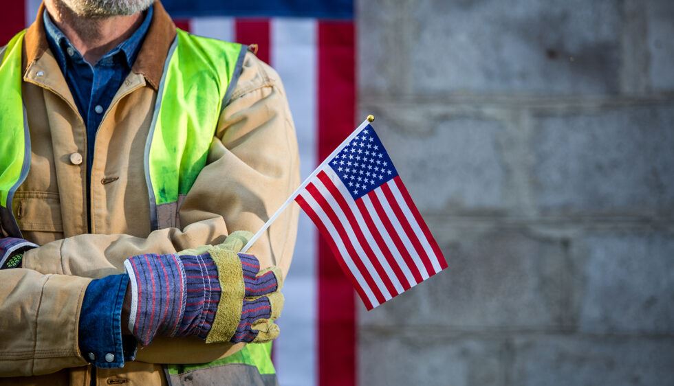 A person wearing a tan jacket, blue shirt, and green safety vest holds a small American flag. The person is also wearing striped work gloves. In the background, part of a large American flag and a gray stone wall are visible.