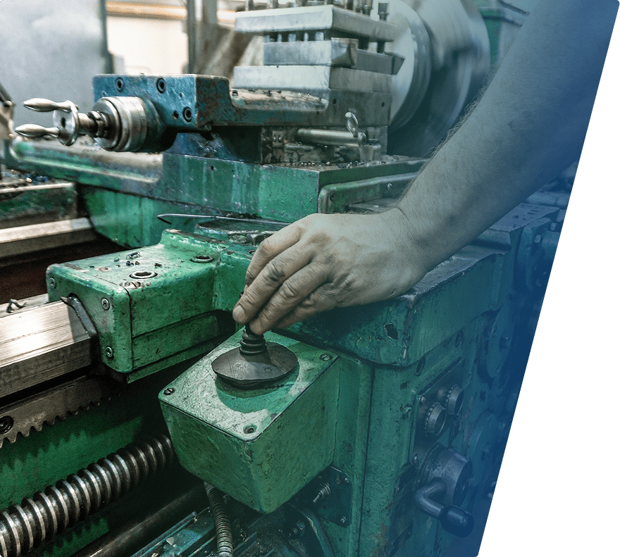 A close-up of a person's hand operating a lever on an old, green industrial lathe machine. The machine shows signs of wear and rust, with various metal components and adjustment knobs visible. The background is slightly blurred, emphasizing the hand and the lever.