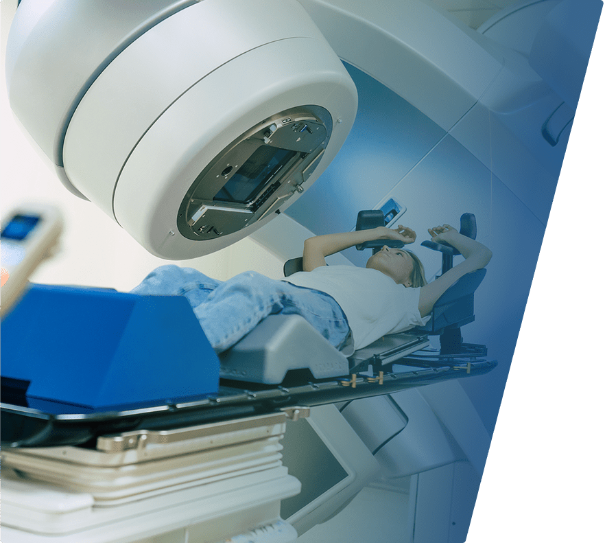 A person lies on a treatment table under a large, circular medical machine, likely a radiation therapy device, in a clinical setting. The individual is positioned with arms raised and appears to be undergoing a medical procedure. The environment is clean and modern, with blue and white tones.