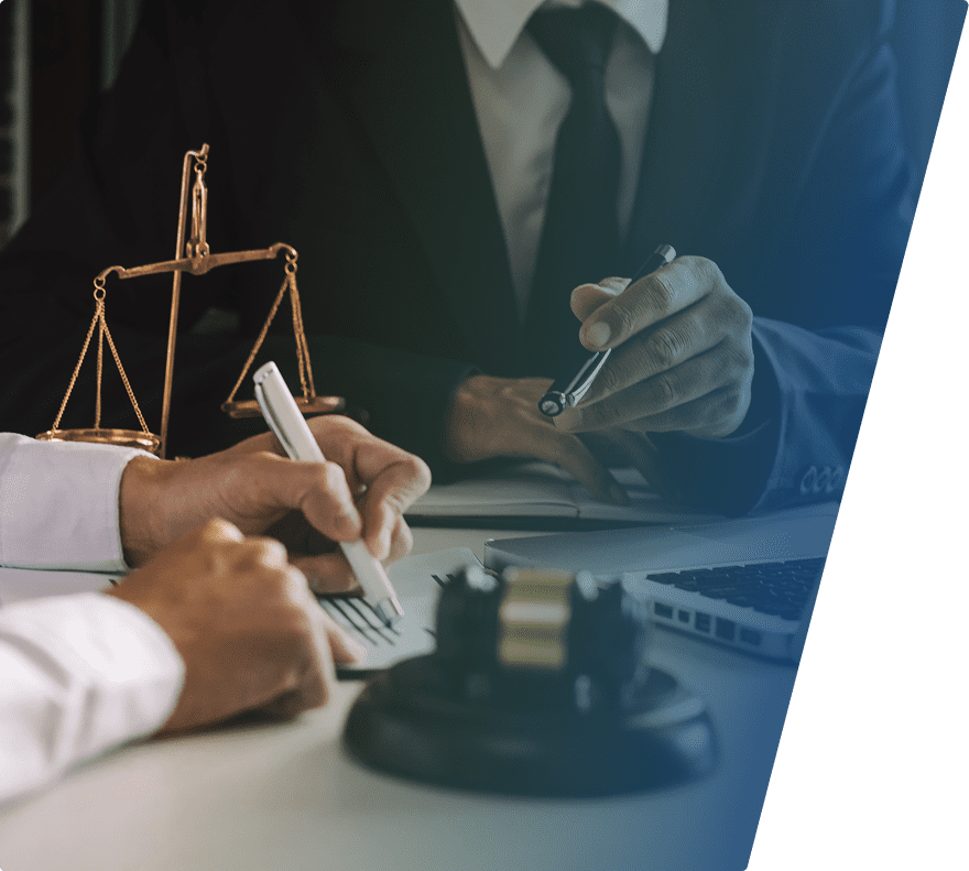 Two people in formal attire are engaged in a discussion or consultation, with one person writing on a document and the other holding a pen. A set of brass scales and a judge's gavel are visible on the table, along with a laptop, suggesting a legal or professional setting.