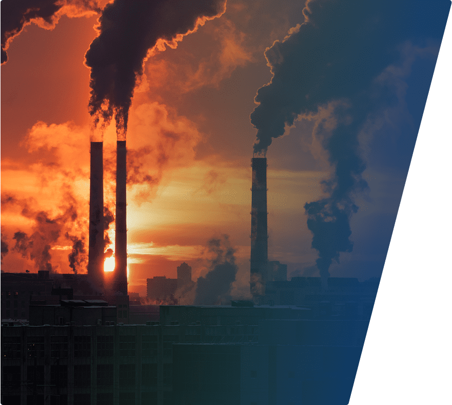 Four industrial smokestacks emitting thick black smoke against an orange sunset sky, with factory buildings silhouetted in the foreground.