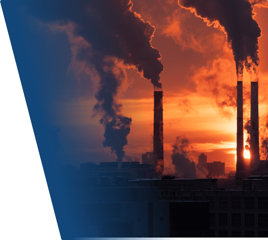 Smoke stacks emitting thick dark smoke against an orange sunset sky, with industrial buildings silhouetted in the foreground.