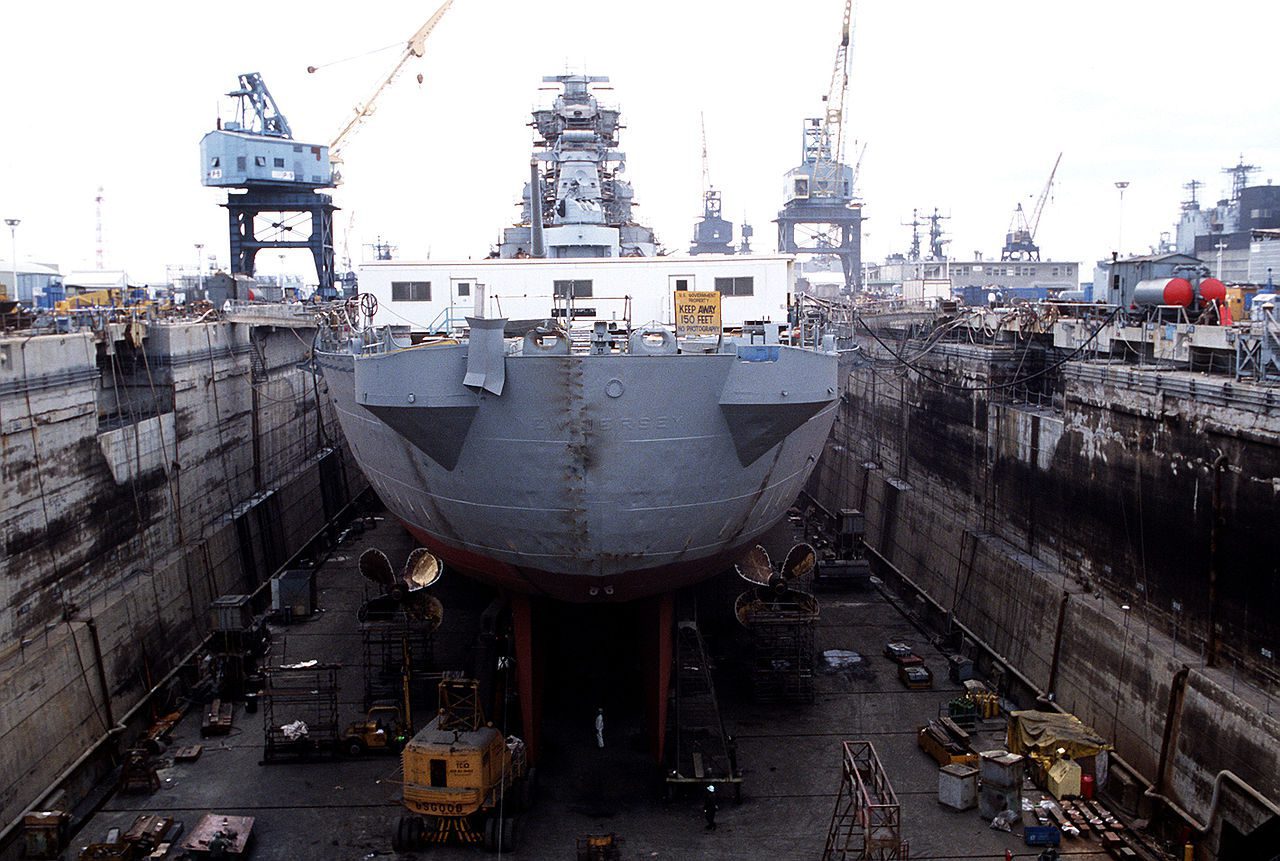 A large naval ship is docked in a dry dock for maintenance or repairs. The ship's hull is visible, with propellers exposed on either side near the stern. The dry dock is surrounded by tall concrete walls, and various equipment, scaffolding, and machinery are scattered around the dock floor. Several cranes and other ships are visible in the background. A sign on the ship reads "KEEP AWAY 150 FEET NO PHOTOGRAPHY.