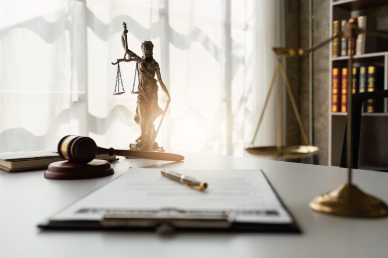 Bronze statue of Lady Justice holding scales and a sword, a wooden gavel on a sound block, a pen on a clipboard with documents, and a set of golden scales on a white desk in a law office with bookshelves in the background.